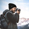 man with camera and backpack wearing black Finvizo beanie in snowy mountains